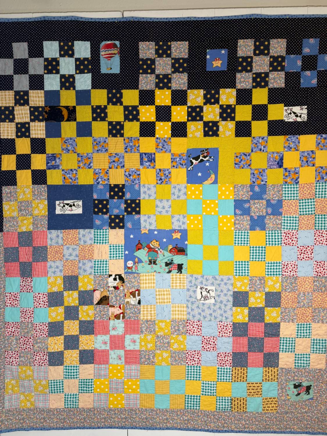 A nine-patch quilt that moves from a starry navy sky at the top to bright gingham and calico below, with nursery-rhyme pictures set in among the squares: a cow jumping over the moon, a hot-air balloon, a cat with a fiddle