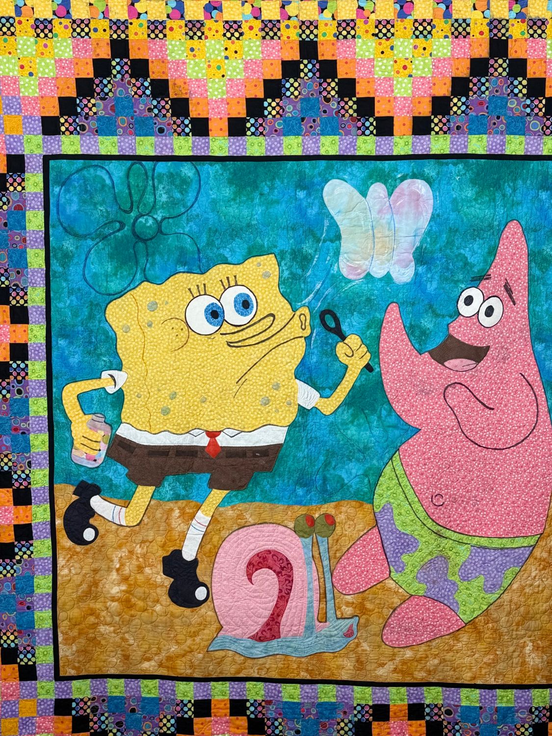 An appliquéd quilt of SpongeBob blowing bubbles beside Patrick and Gary the snail on a mottled blue sea, framed by a border of bright polka-dot squares