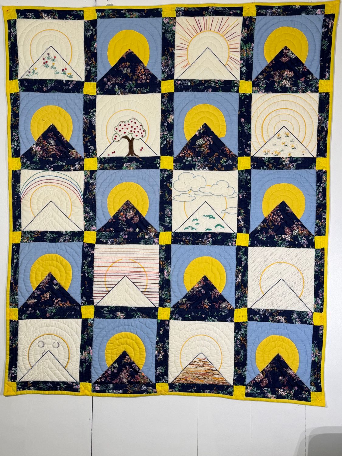 A quilt of twenty blocks, each a triangle mountain under a rising sun — appliquéd yellow suns on blue alternating with embroidered blocks of flowers, a tree, a rainbow and clouds — in navy floral sashing with yellow cornerstones